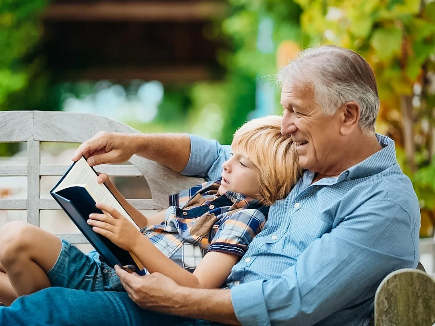 A grandfather reading aloud to his grandson from a dark hardcover on a garden bench