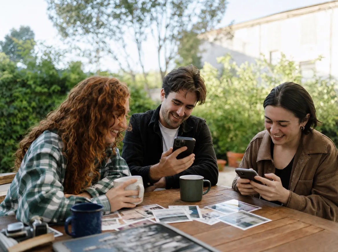 Three friends at a garden table comparing travel photos on their phones