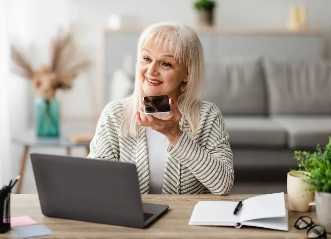 An older woman speaks her answer into her smartphone, laptop and notebook on the table