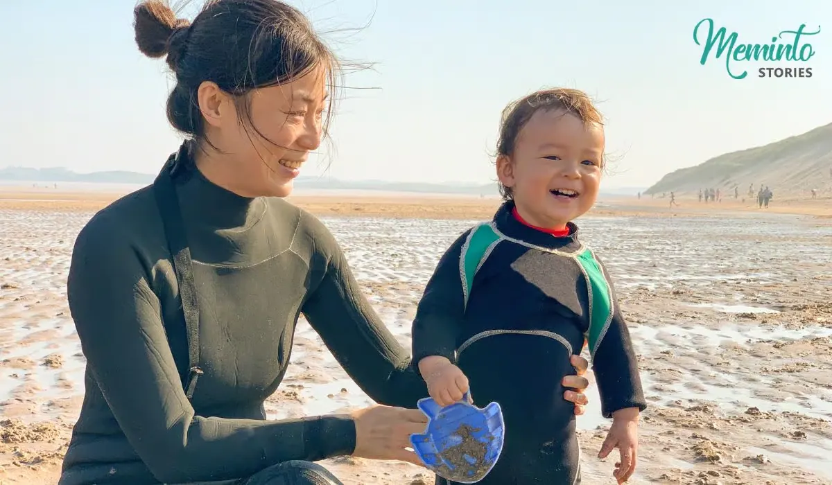 Mutter und lachendes Kleinkind im Neoprenanzug hocken gemeinsam am Strand