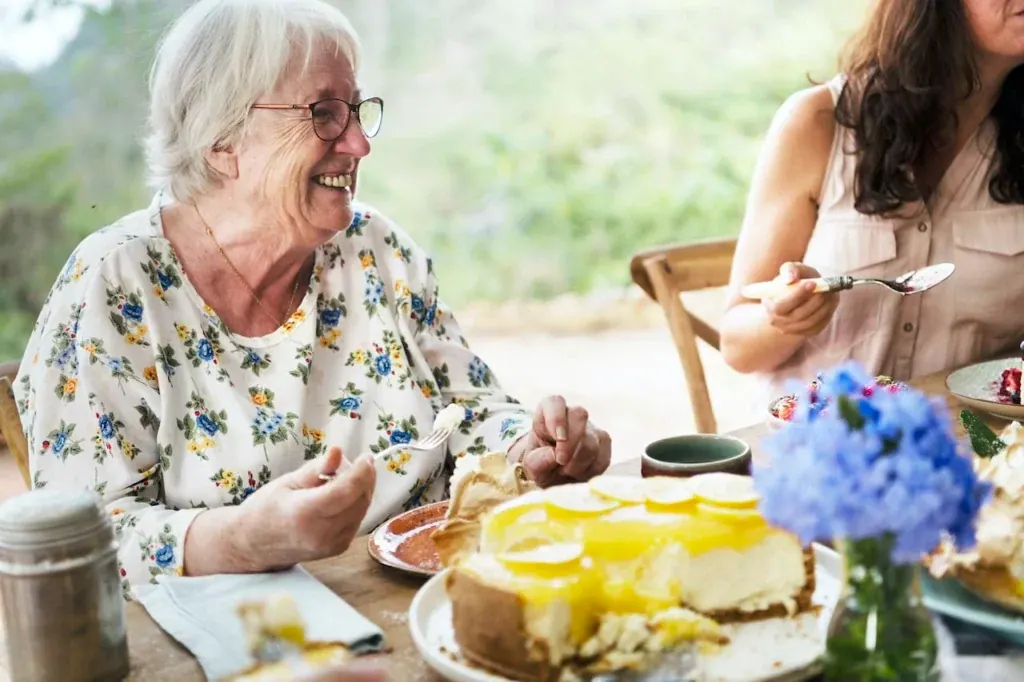Lachende Seniorin an einer sommerlichen Kaffeetafel im Garten, vor ihr ein Zitronenkuchen