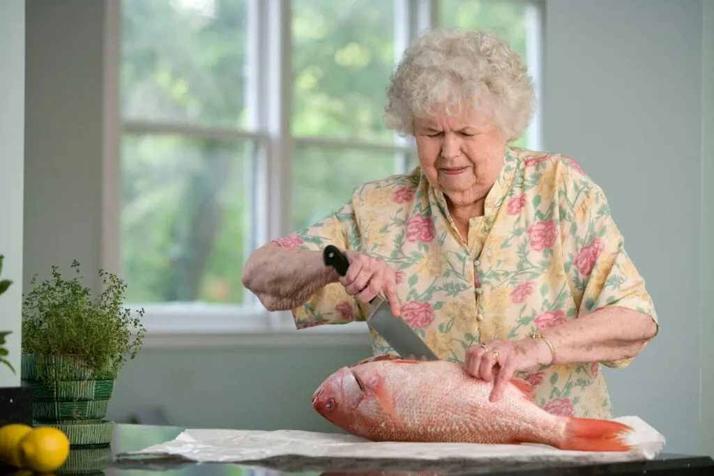 Older woman in a floral blouse preparing a whole fish at her kitchen counter